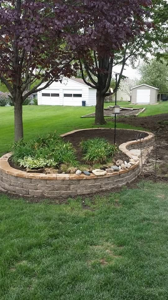 Curved block raised garden bed — backyard with mature trees and outbuildings