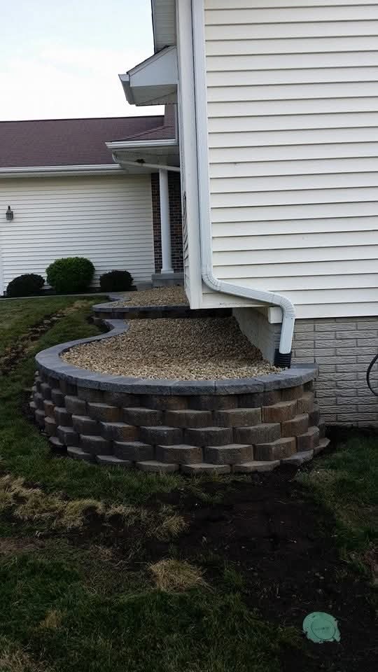 Curved block retaining wall with gravel bed — side of home with downspout